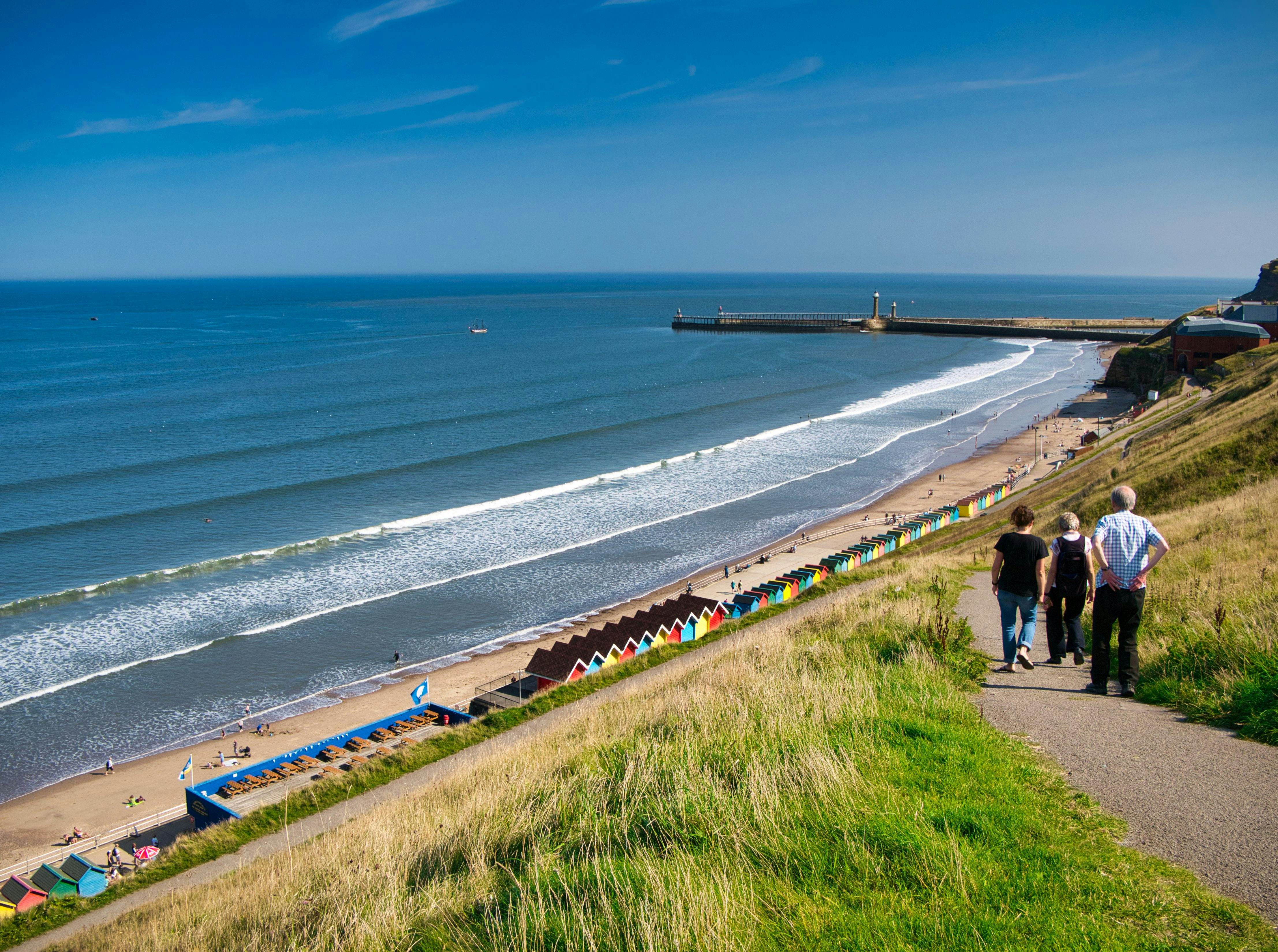 Tourists on a path overlooking colourful beach huts, the coast and harbour piers at Whitby, North Yorkshire, UK - taken on a sunny day at the end of summer